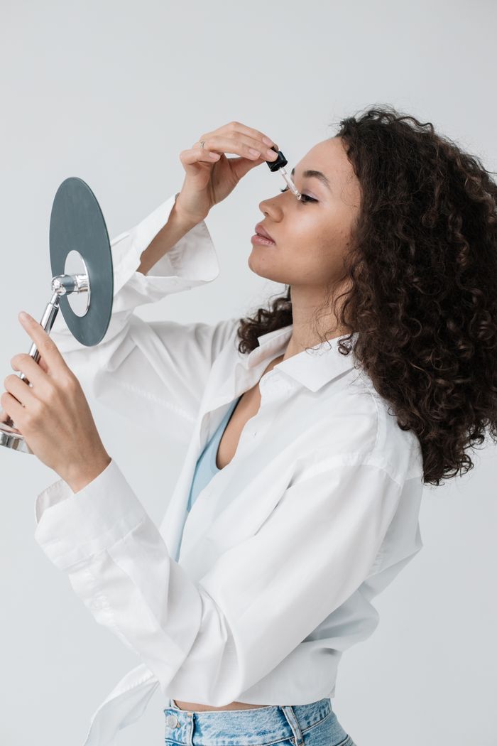 services-01 A woman with curly hair applies skincare serum using a dropper in front of a mirror.